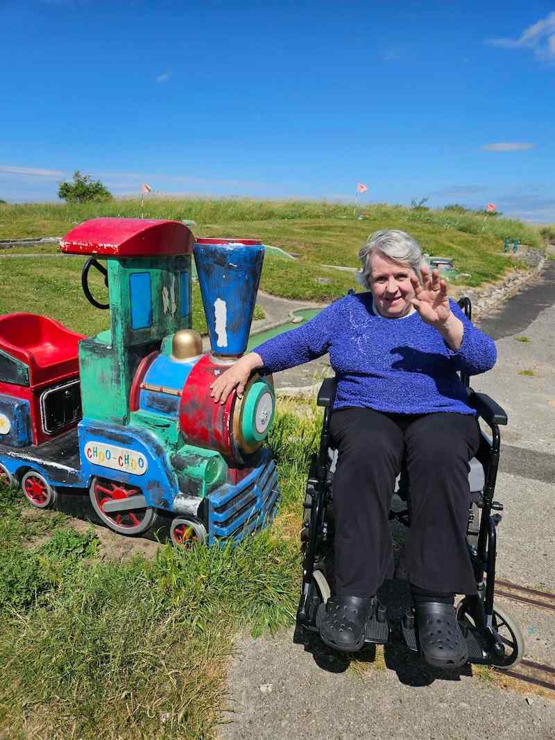 elderly female sat in a wheelchair in the local community next to a model train smiling