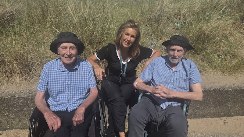2 male residents sat in wheelchairs smiling with a carer outside